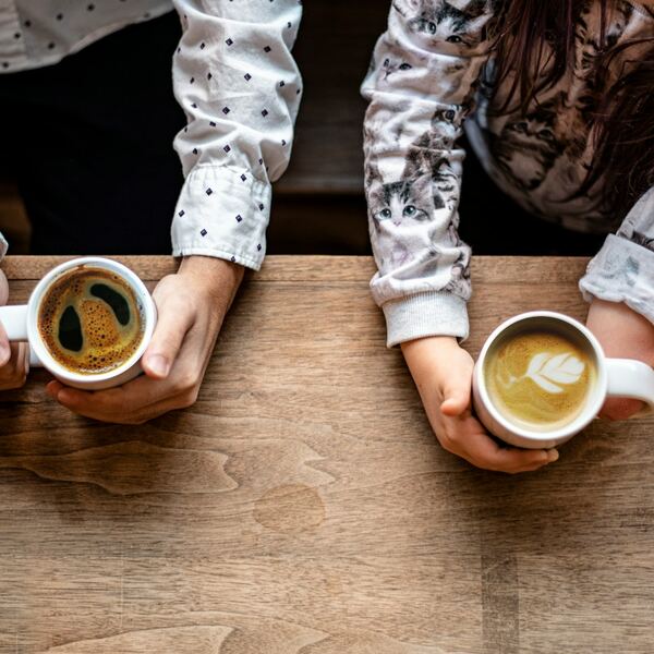 Twee paar handen houden een kop koffie vast aan weerszijden van een eikenhouten tafel, van bovenaf gezien.